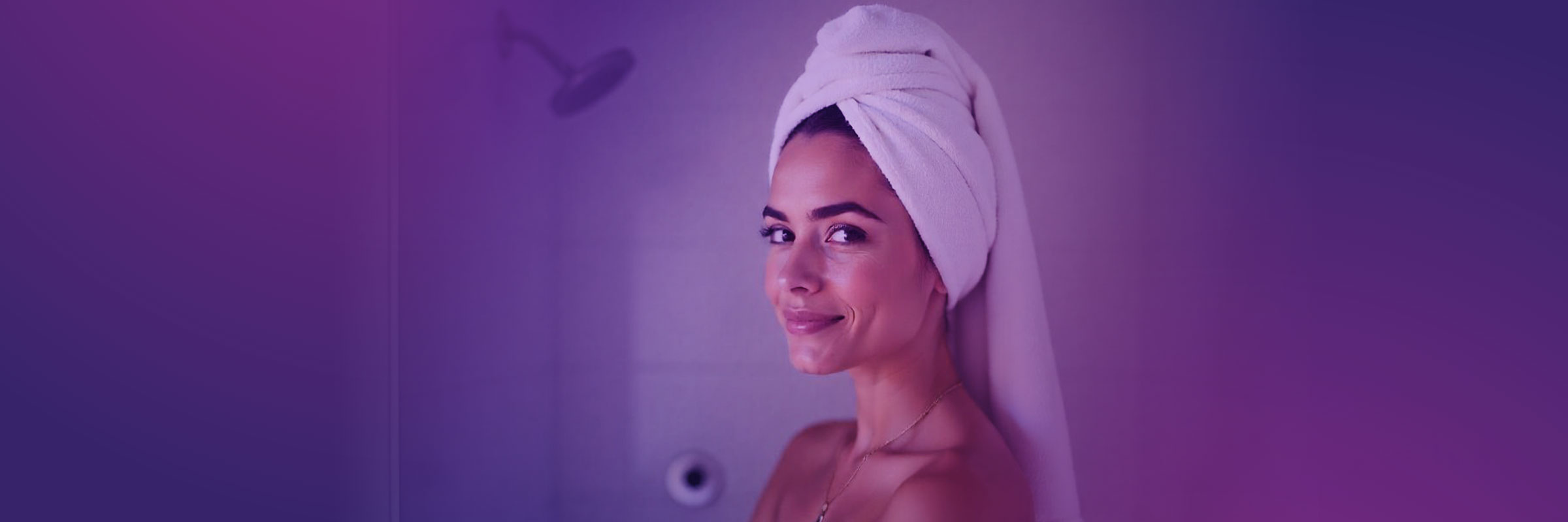 A person stands in a softly lit shower, wrapped in a white towel around their head, smiling slightly. The background has a smooth gradient of purple hues, creating a calm and serene atmosphere.