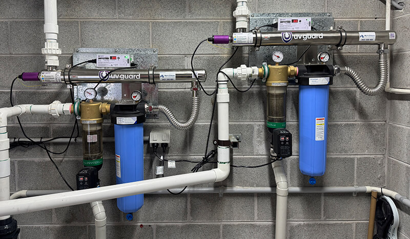 A water filtration system setup against a gray brick wall, with two UV-guard units and connected pipes. Each unit has a gauge, electrical connections, and blue filter housings.