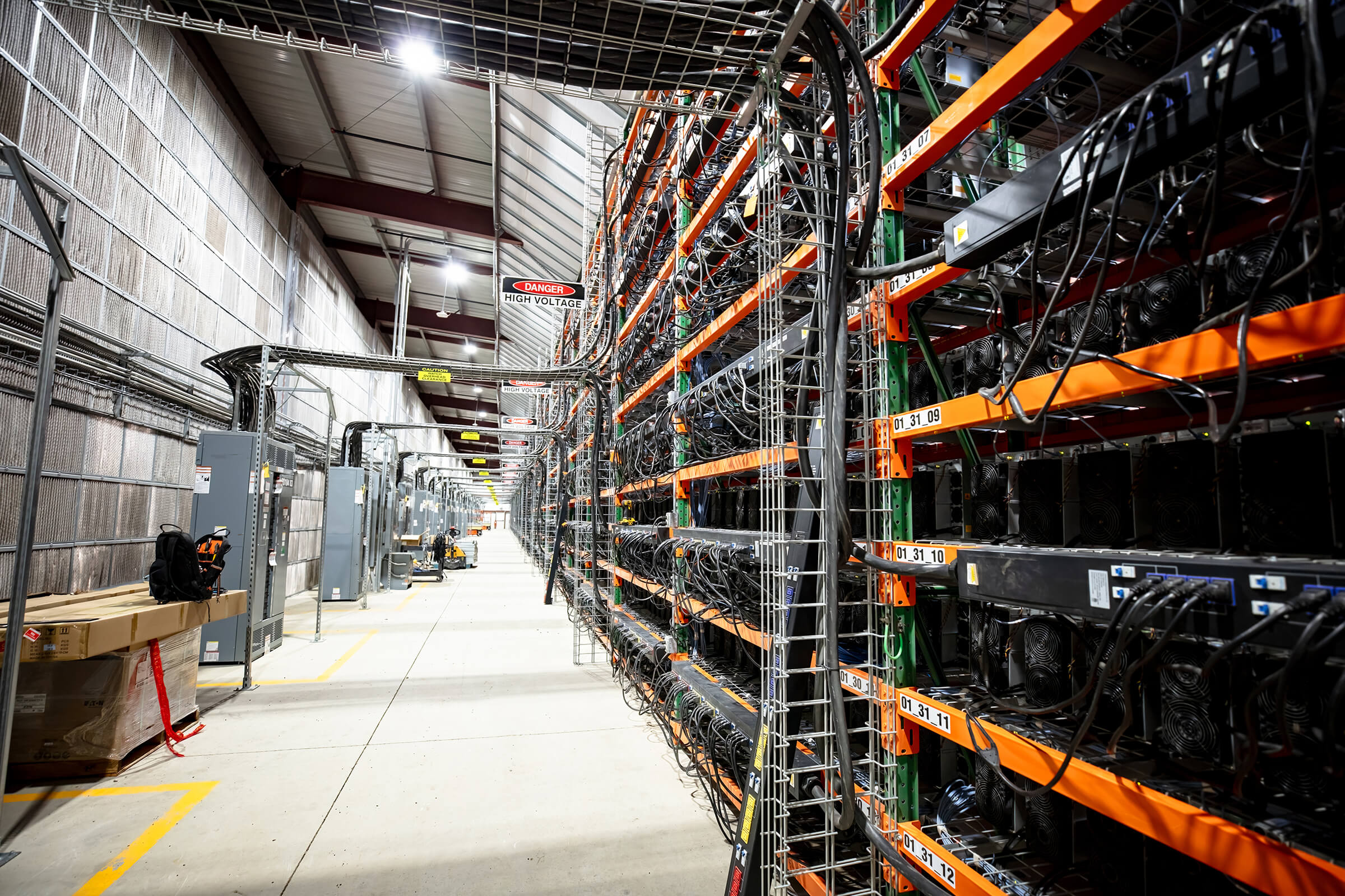 A large industrial facility with rows of computer servers and cables on metal racks, used for cryptocurrency mining. The space is well-lit and organized, with concrete floors and high ceilings. A large industrial facility with rows of computer servers and cables on metal racks, used for cryptocurrency mining. The space is well-lit and organized, with concrete floors and high ceilings.