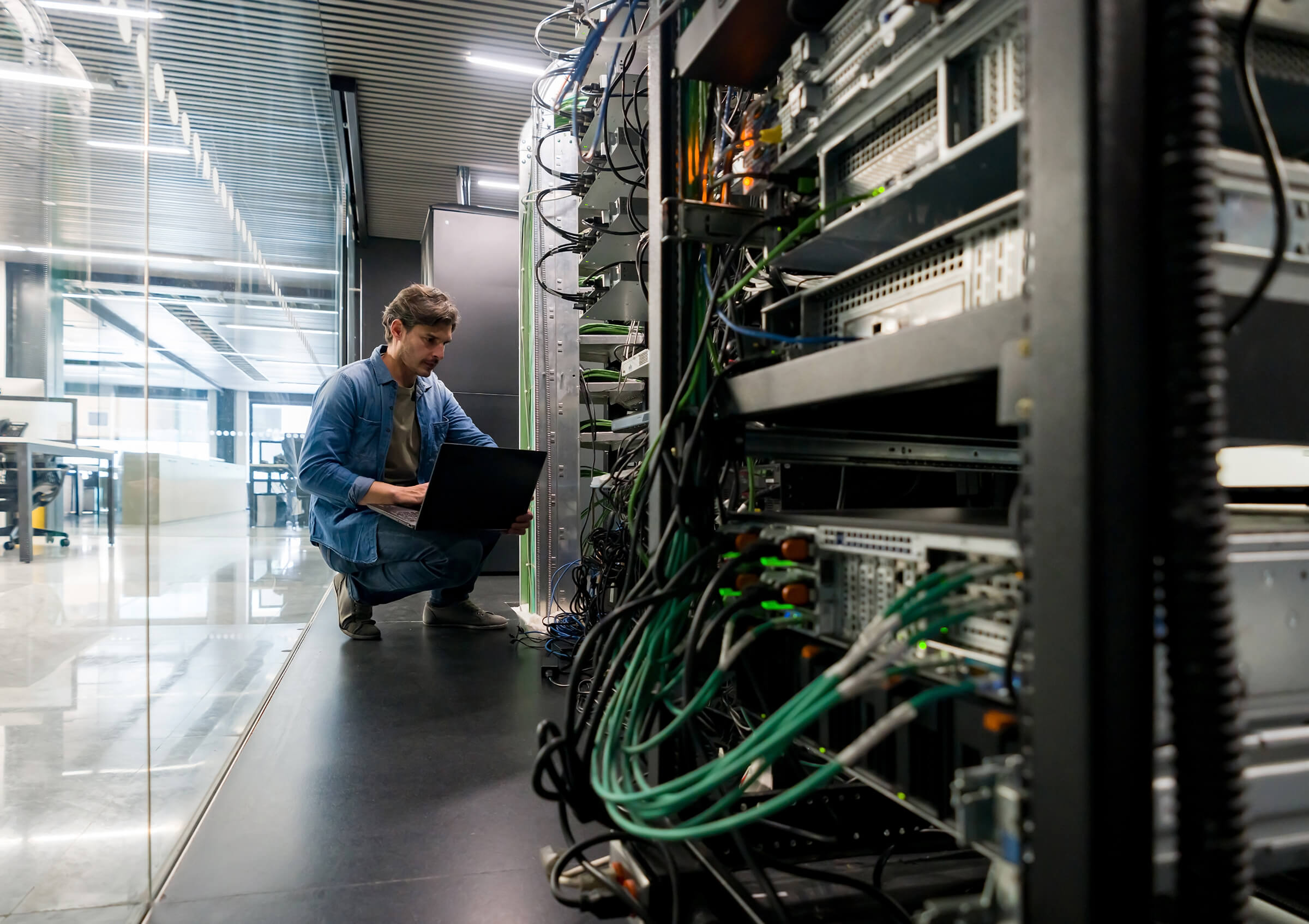 A man kneels on the floor in a modern server room, working on a laptop beside several server racks with visible cables and wires. The space appears clean and professional. A man kneels on the floor in a modern server room, working on a laptop beside several server racks with visible cables and wires. The space appears clean and professional.